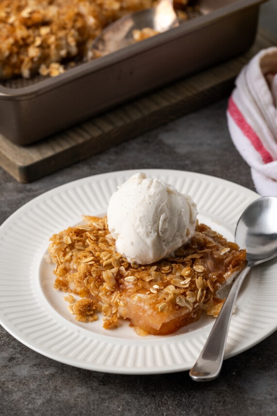 A serving of apple dump cake on a white plate topped with a scoop of ice cream, next to a spoon.