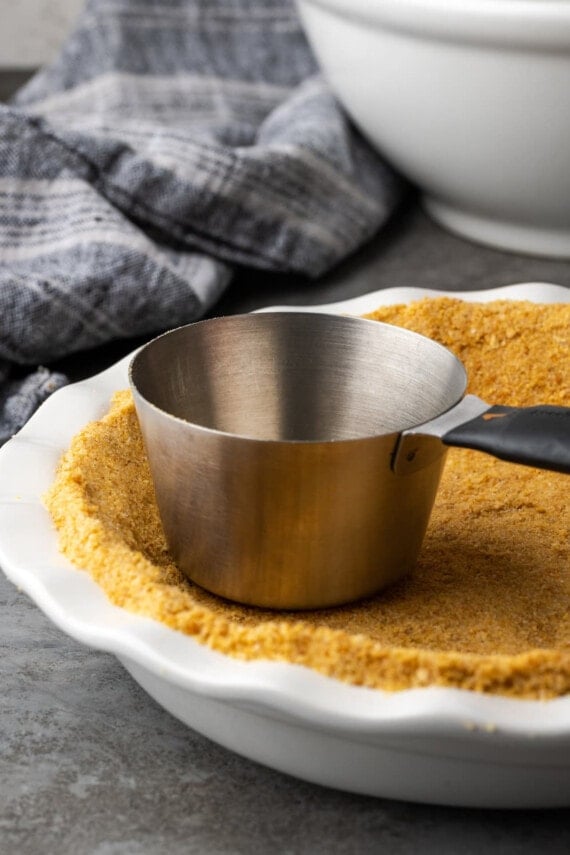 A metal measuring cup is used to press the graham cracker crust into a white pie dish.