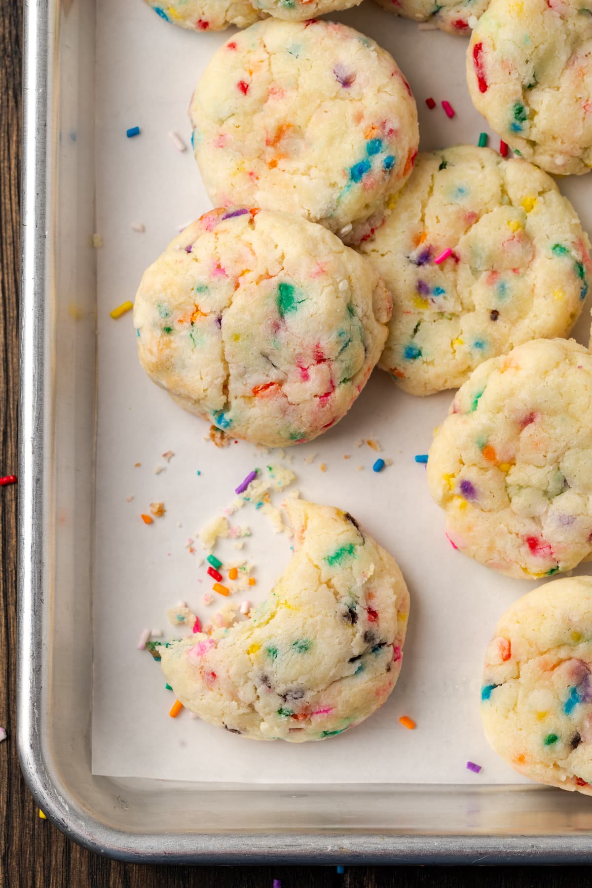 Close up overhead view of assorted funfetti cookies on a parchment-lined baking sheet, with a bite missing from one cookie.