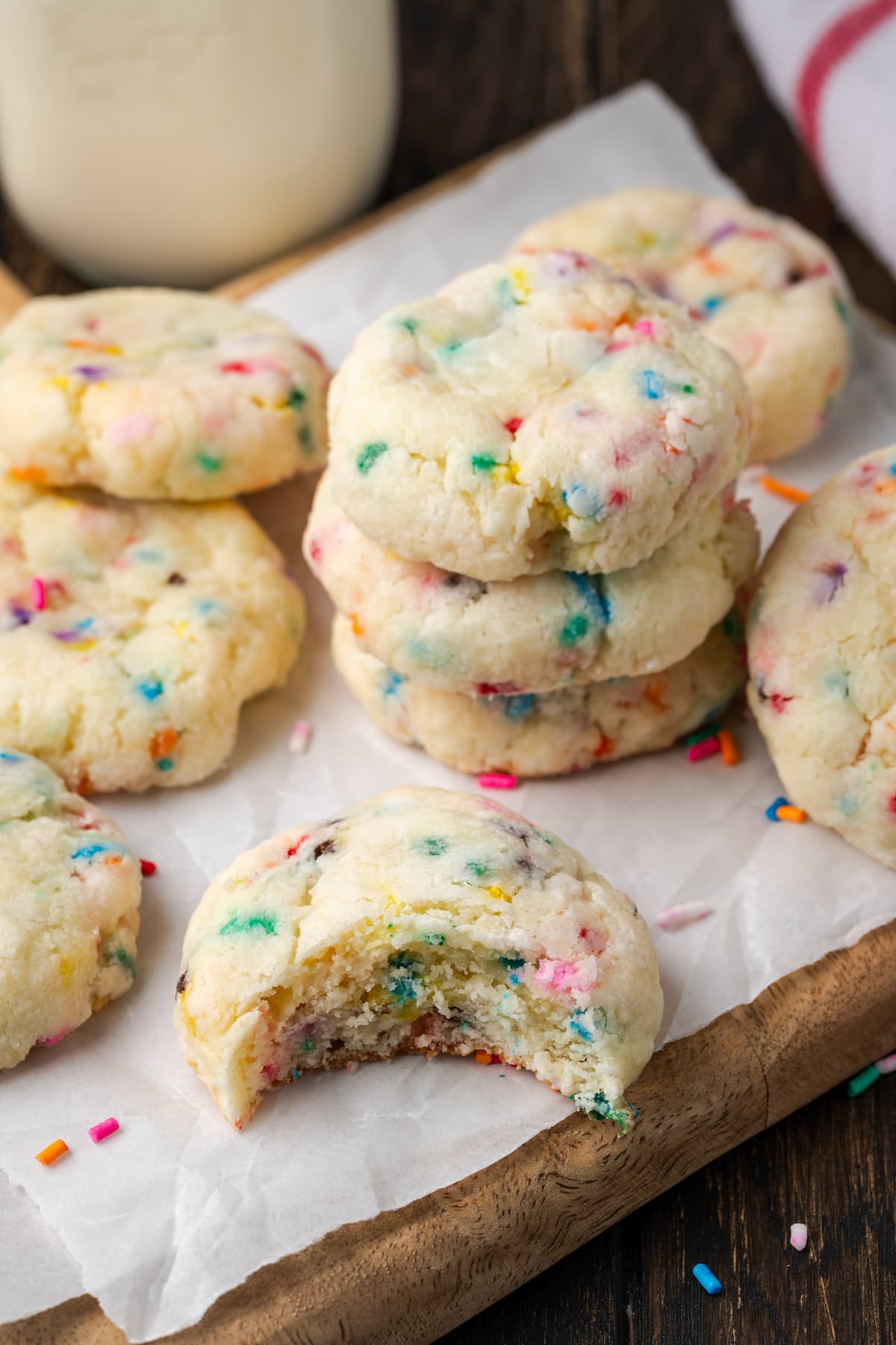 Stacks of funfetti cookies on a parchment-lined wooden cutting board, next to one cookie with a bite missing.