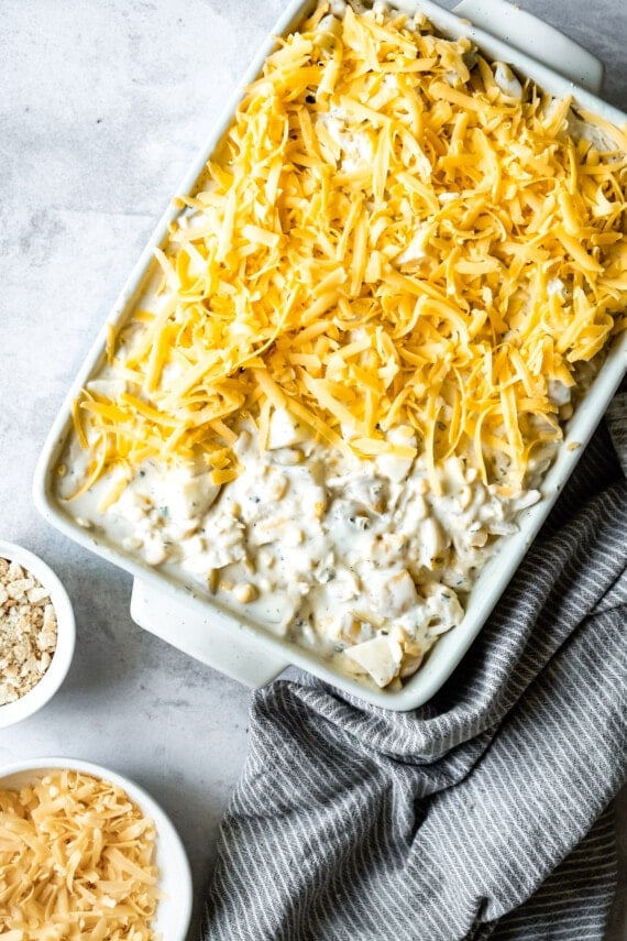 An unbaked chicken hashbrown casserole in a baking dish partially topped with shredded cheese.