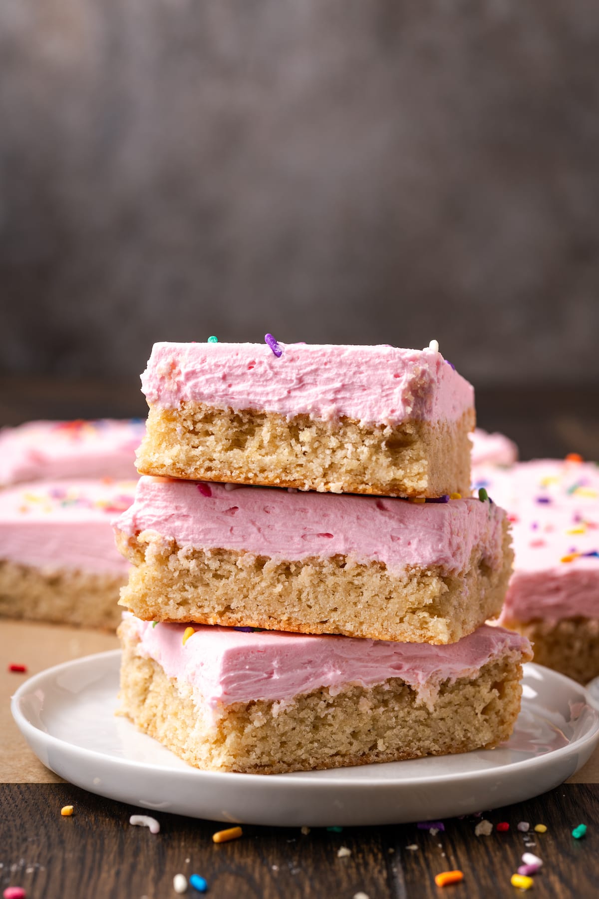 Three frosted sugar cookie bars stacked on a white plate, with more bars in the background.