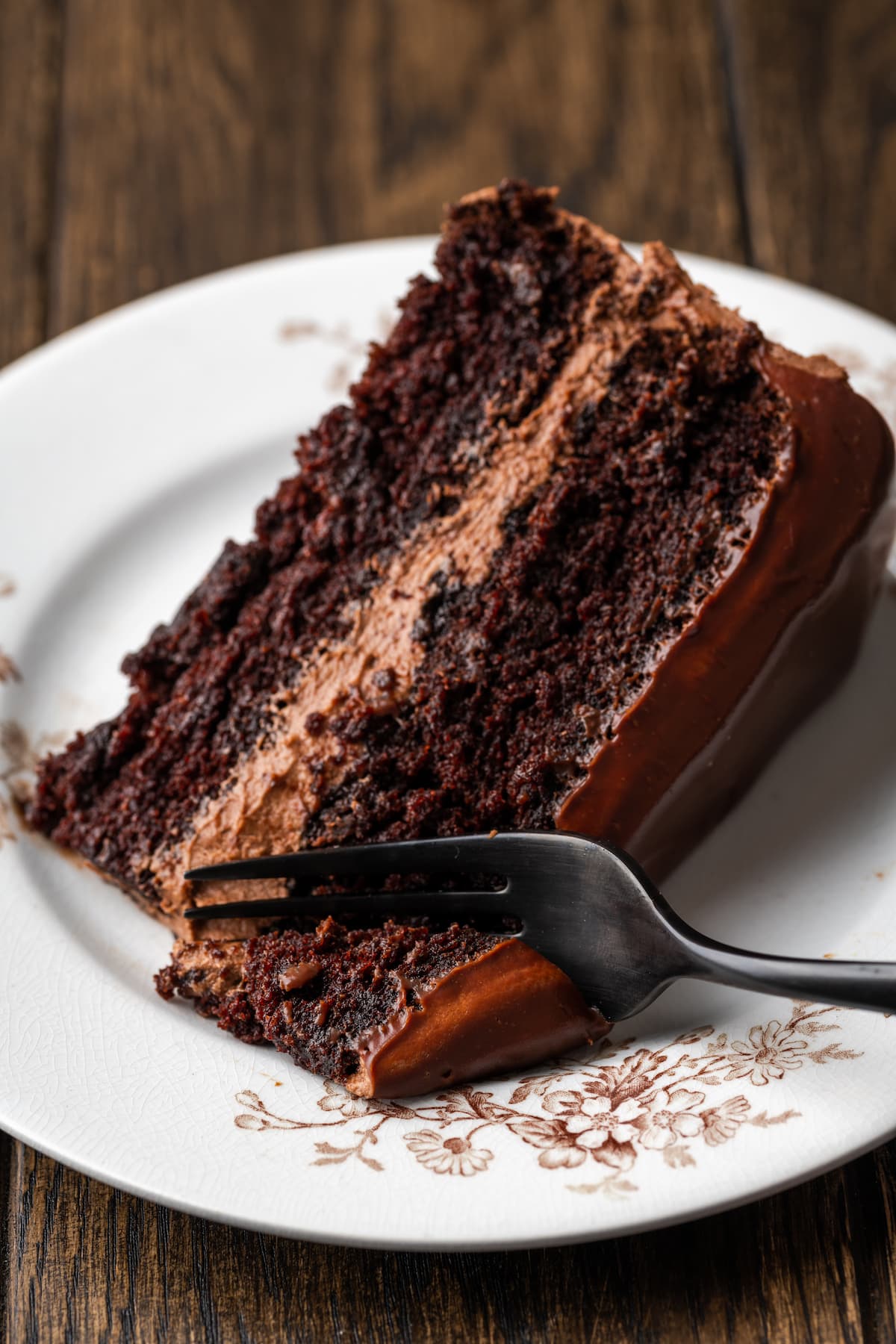 A forkful of chocolate cake next to a slice of chocolate layer cake on a white plate.