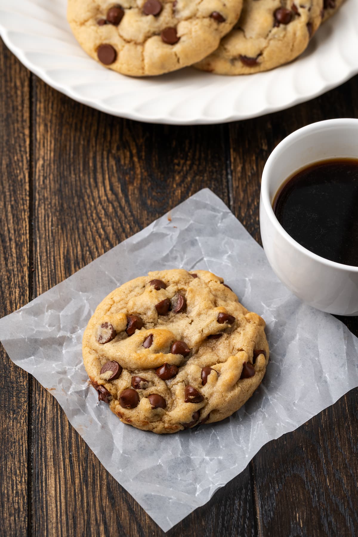 A chocolate chip Subway cookie on a piece of parchment paper next to a cup of coffee, with more cookies on a white plate in the background.