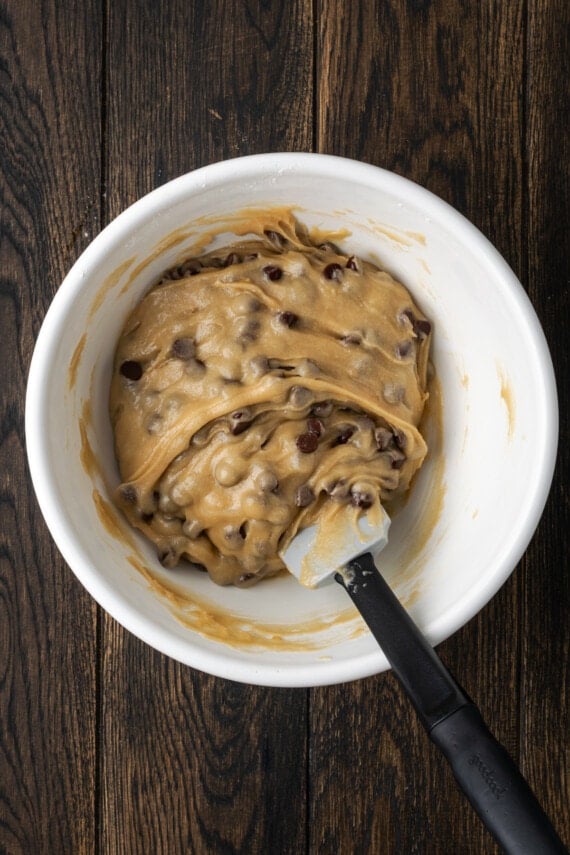 Chocolate chips folded into cookie dough in a white bowl, with a spatula.