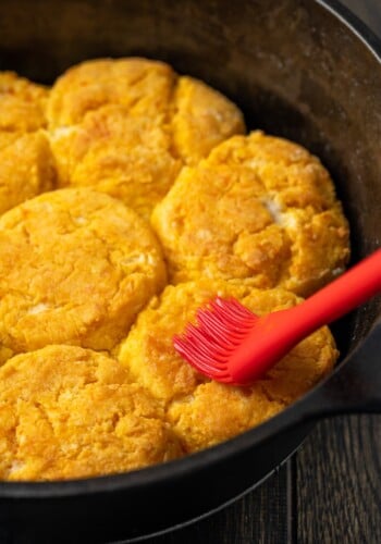A rubber spatula brushing melted butter over baked sweet potato biscuits in a cast iron skillet.
