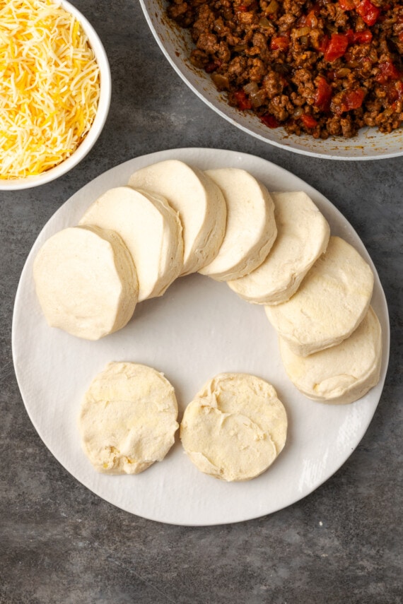 Biscuit dough discs on a white plate next to bowls of taco meat and shredded cheese.