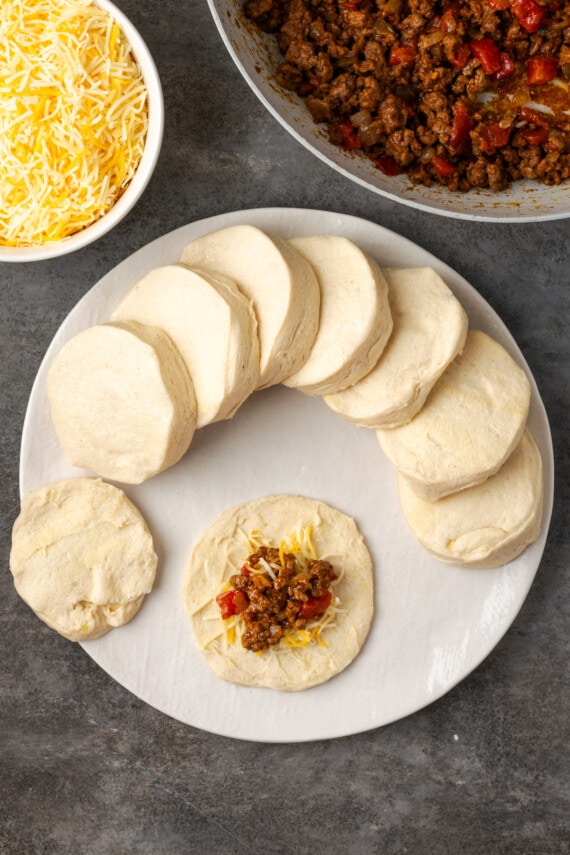 Ground taco meat added over top shredded cheese on flattened biscuit dough, next to more biscuit dough discs and bowls of taco meat and shredded cheese.